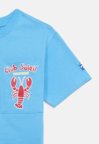 Μπλε πουκάμισο με κοντά μανίκια και μπροστινή τσέπη που φέρει ένα κόκκινο σχέδιο αστακού και το κείμενο "Club Soleil Good Vibes" σε κόκκινο και κίτρινο.