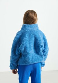 Chaqueta de forro polar azul suave con cuello alto y corte relajado. El material tiene una superficie suave y texturizada. Llevada con pantalones azules a juego.