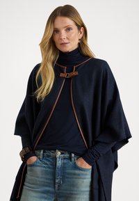 Jūrinės vilnos pončas su rudos odos akcentais, turintis priekinį sagties uždėjimą ir laisvas trumpas rankoves. Pora su tamsiu golfu ir šviesiomis džinsomis.