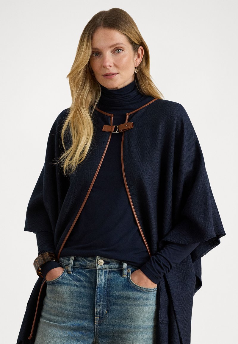 Jūrinės vilnos pončas su rudos odos akcentais, turintis priekinį sagties uždėjimą ir laisvas trumpas rankoves. Pora su tamsiu golfu ir šviesiomis džinsomis.