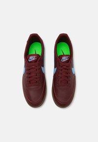 Nike copalke iz burgundskega usnja z svetlo modrimi detajli. Značilnosti vključujejo okroglo konico, ravne vezalke in zelene notranjosti s logotipom.