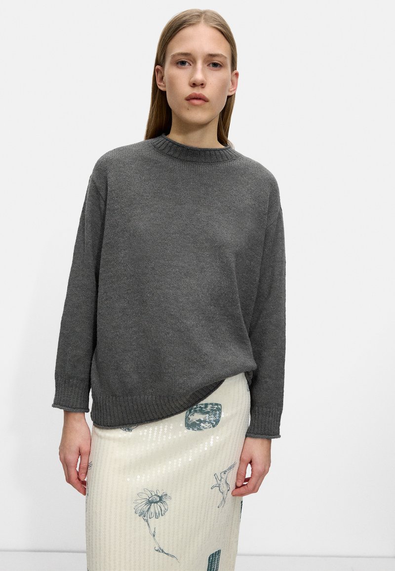 Szary dziergany sweter z okrągłym dekoltem i długimi rękawami, w połączeniu z kremową spódnicą z cekinami, zdobioną zielonymi kwiatowymi i geometrycznymi wzorami.