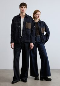 Μπουφάν από μαύρο τζιν με καφέ τσέπη, φορεμένο πάνω από λευκό αμάνικο τοπ; συνδυασμένο με φαρδιά, σκούρα τζιν παντελόνια. Και οι δύο φορούν μαύρα παπούτσια, στέκονται μπροστά σε ουδέτερο φόντο.