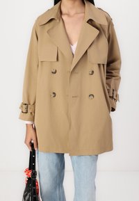 Beige dubbelzijdige trenchcoat met grote kraag, knoopaccenten en riem manchetten; gedragen over een lichte top en gecombineerd met lichte jeans.
