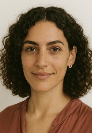 Ritratto di una persona con capelli ricci e scuri, che indossa una maglietta color ruggine, con lentiggini sottili e piccoli orecchini rotondi, su uno sfondo neutro.