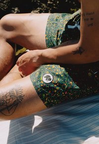 Zelena kupaćica s uzorkom koja sadrži šarene ilustracije, okruglu oznaku logotipa i širok kroj. Vidljive tetovaže na ruci.