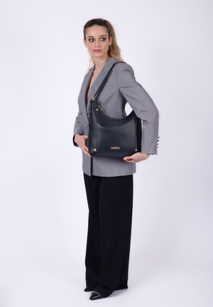Kvinde i grå blazer og sorte bukser, der holder en stor sort håndtaske over sin skulder, stående mod en ensfarvet hvid baggrund.