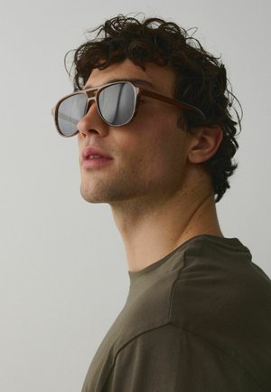 Junger Mann mit lockigem Haar, der braun gerahmte Aviator-Sonnenbrillen trägt und ein olivgrünes Hemd anhat, schaut leicht nach oben vor einem einfarbigen Hintergrund.