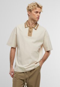 Beige Poloshirt mit kurzen Ärmeln und einem strukturierten braunen Kragen, der zwei braune Knöpfe und weiße Streifen aufweist. Dezentes Logo an der Seite.