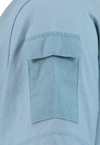 Helles blaues Stoff mit einer genähten Klappen-Tasche an einem Kleidungsstück, das detaillierte Nähte und Textur im Ärmelbereich zeigt.