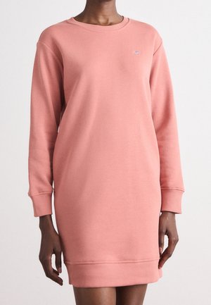 Personne portant une robe sweat-shirt rose poudré à manches longues, longueur genou, avec poignets et ourlet côtelés, sur un fond uni.
