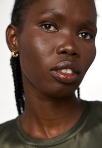 Gros plan sur un visage avec une peau lisse, des yeux marron foncé et des cheveux tressés. Porte un vêtement vert et de petites boucles d'oreilles en or.