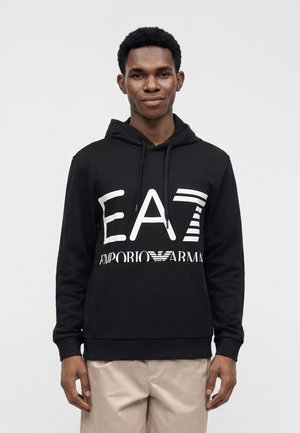 Jeune homme portant un sweat à capuche noir avec logo blanc EA7 Emporio Armani, pantalon beige, debout devant un fond clair uni.