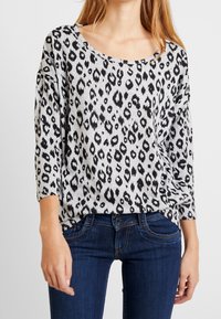 Haut à manches longues gris avec un motif léopard noir. Doté d'un décolleté large et d'une coupe décontractée, associé à un jean bleu foncé.