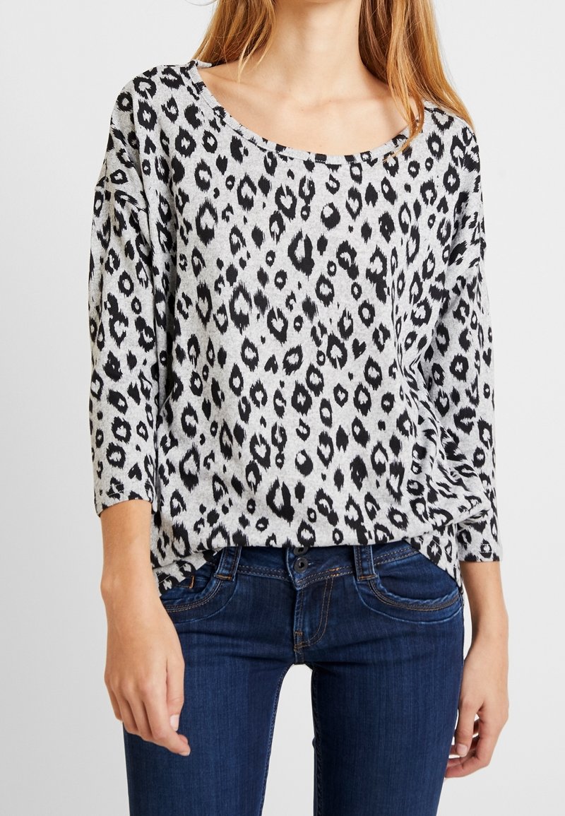 Haut à manches longues gris avec un motif léopard noir. Doté d'un décolleté large et d'une coupe décontractée, associé à un jean bleu foncé.