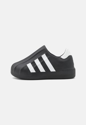 Housse de chaussure noire slip-on avec des bandes blanches de style Adidas et une languette blanche au talon sur un fond clair uni.