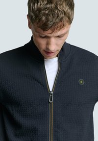 Zwarte zip-up jacket met een gestructureerd patroon, voorzien van een ritssluiting en een hexagonaal logo accent. Een wit t-shirt eronder is zichtbaar.
