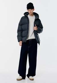 Kurtka puffer w ciemnoszarym kolorze, z kapturem, zapięciem na zamek i pikowanym wzorem; zestawiona z luźnymi, ciemnoniebieskimi dżinsami i białymi trampkami.