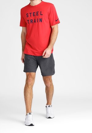 Uomo che indossa una maglietta rossa "STEEL TRAIN", pantaloni corti grigio scuro e sneakers bianche Nike, in piedi contro uno sfondo chiaro e uniforme.