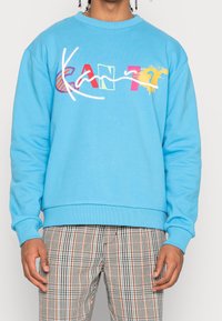 Felpa in cotone blu con design del testo colorato "CAN?" e dettagli bianchi. Presenta un colletto a girocollo e polsini e orlo a coste. Pantaloni a quadri in basso.