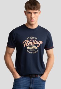 Tums tumši zils kokvilnas t-krekls ar retro stila krēmkrāsas un sarkana grafiku, kurā ir vārds "Heritage", papildināts ar kontrastējošām sarkanām šuvēm uz piedurknēm.