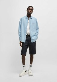 Ljusblå jeansskjorta medknappar, vit T-shirt, mörka shorts, vita sneakers med svarta detaljer och logosockor. Modellen står med händerna i fickorna.