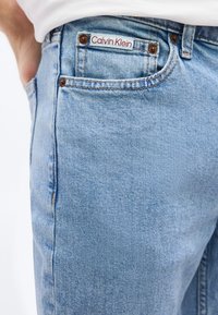 Τσέπη από ανοιχτού μπλε τζιν με λευκή και κόκκινη ετικέτα Calvin Klein και εμφανείς χαλκοκονδυλιές, φορεμένη με λευκό τοπ.