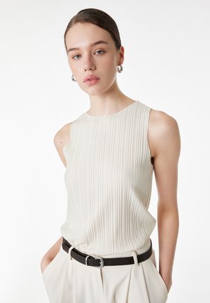 Jeune femme aux cheveux lissés en arrière portant des boucles d'oreilles argentées en forme d'anneaux, un haut plissé sans manches blanc, un pantalon blanc et une ceinture noire, les mains dans les poches.