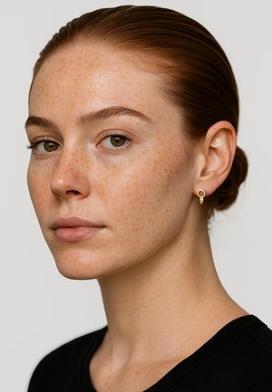 Goldene Creolen mit kreisförmigem Design, getragen von einem Modell mit heller Haut und Sommersprossen, zusammen mit einem schlichten schwarzen Shirt.