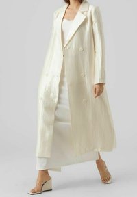 Dubbelknäppt, lång kappa i iriserande krämfärgad tyg, med spetsig krage och framsida knappar, lager över en vit klänning och nakna klackar.