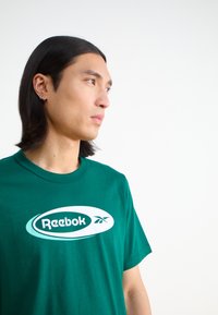 Žalia medvilninė marškinėliai su dideliu baltu Reebok logotipu, apvaliu iškirpimu ir trumpomis rankovėmis, pasižymintys lygia tekstūra.
