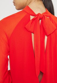 Punainen pusero, jossa on korkea kaulus, avonainen selkäosa ja jousirinne suljenta niskassa. Kangas vaikuttaa sileältä ja kevyeltä.