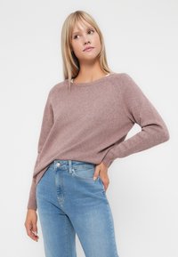 Lys rosa strikkegenser med rund hals og ribbestrikkede mansjetter, kombinert med blå høy midje jeans, stående mot en ensfarget bakgrunn.