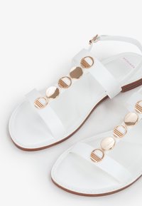 White flat sandals with multiple gold circular accents and two strap design. Made of smooth synthetic material with a brown sole.