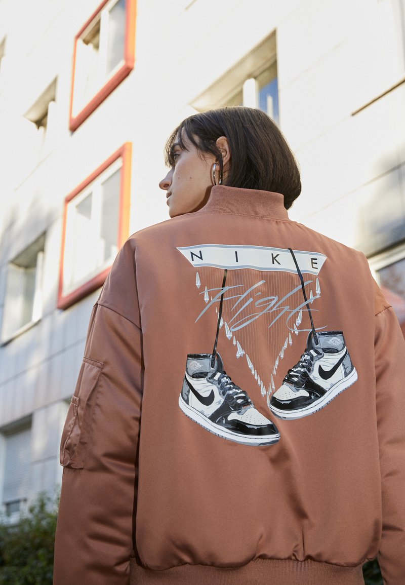 Veste bomber Nike en tissu marron, avec un graphique de baskets au dos accompagné du texte "FLIGHT", col montant et taille élastique.