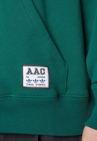Grön sweatshirt med texturerad tyg. Har en broderad AAC by adidas-logotyp och Tre Stripes-logotyp. Regular fit-design med en känguruficka.