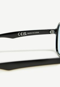 Fekete napszemüveg téglalap alakú kerettel, fényes felülettel. A szárakon a "CE UKCA MADE IN CHINA" feliratok találhatóak. Átlátszó lencsék.