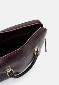 Bordeaux lædertaske med en guldfarvet lynlås, topgreb og en enkelt indre lomme. Glat overflade og rektangulær form.