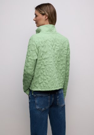 Femme de profil portant une veste texturée vert clair à col haut et un jean bleu en denim, sur un fond uni.