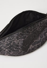 Schwarze und graue gemusterte Nike-Gürteltasche mit einem Reißverschluss; sie hat eine glatte Textur und ein auffälliges schwarzes Logo auf der Vorderseite.