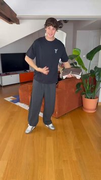 Čierne tričko s logom, šedé tepláky a strieborné tenisky. Drží zložené oblečenie pred textúrovanou oranžovou pohovkou a izbovými rastlinami.