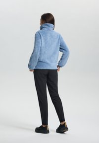 Frau steht mit Händen in den Hüften, trägt eine hellblaue Fleecejacke, schwarze Hosen und schwarze Sneakers mit braunen Akzenten.