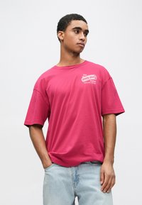 Rosa oversized t-skjorte med korte ermer, med trykt logo på brystet. Kombinert med lyseblå denimjeans.