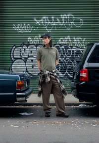 Kaki polo majica, karirasta flanelska srajca zavezana okoli pasu, rjave hlače s črtami, črni supergi. Zelena stena z grafiti, na obeh straneh dve parkirani avtomobili.