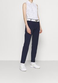 Bílá bezrukávová košile s malými černými tečkami, tmavě námořnické kalhoty a bílé sportovní boty se žlutými akcenty. Obsahuje bílý pás s kovovou sponou.