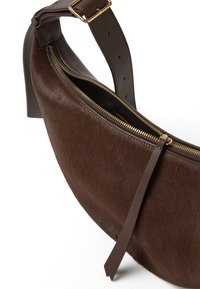 Bolso cruzado de piel marrón con una correa de cuero suave, cremallera en tono dorado y forma plana y curva; cuenta con un cierre de cremallera y un compartimento interior.