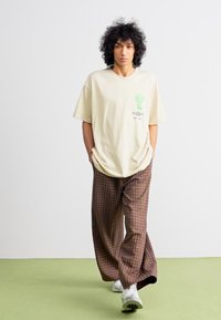 Beige oversized t-skjorte med grønn kaktusgrafikk; kombinert med brune rutete vide bukser. Joggesko fullfører det uformelle utseendet.