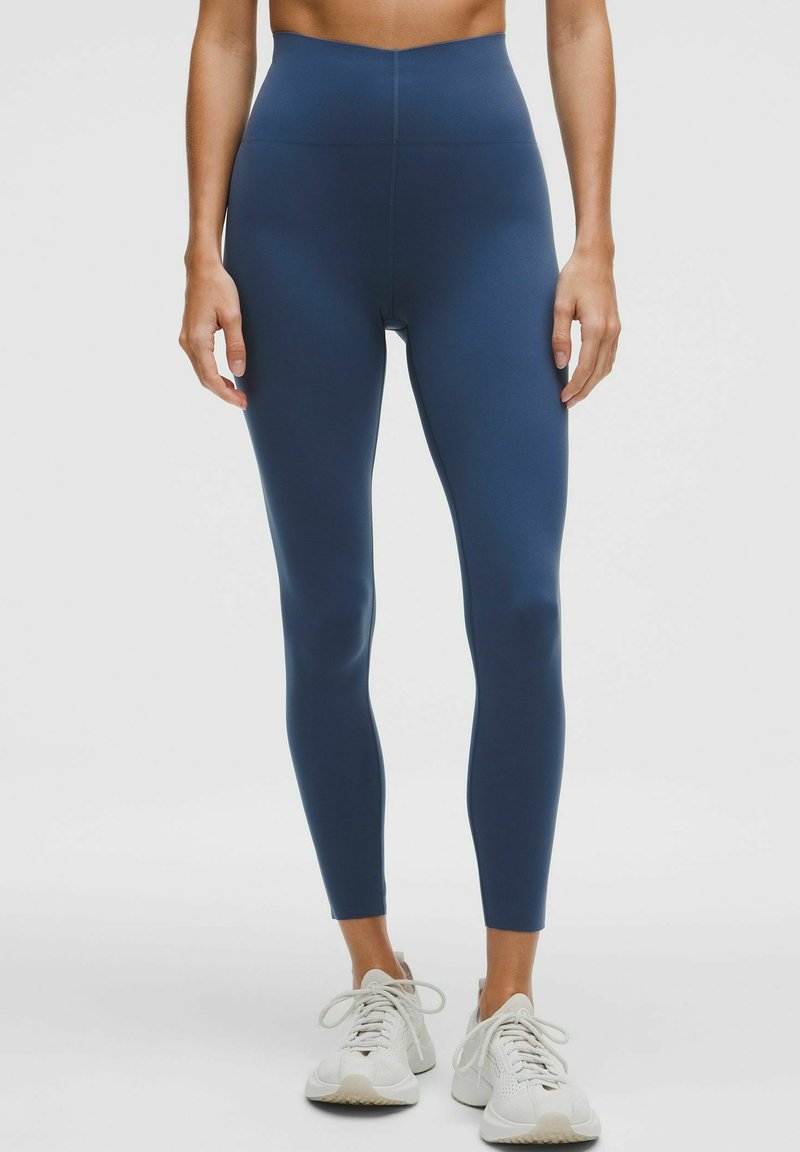 Leggings cu talie înaltă, strâmte, din material elastic de culoare albastru închis, cu o textură netedă, purtate împreună cu adidași de culoare gri deschis.