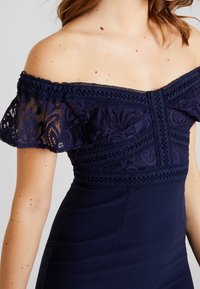 Marinblå klänning med avtagna axlar som har en spetsöverdel, intrikata mönster och en figurnära silhuett, slät textur.