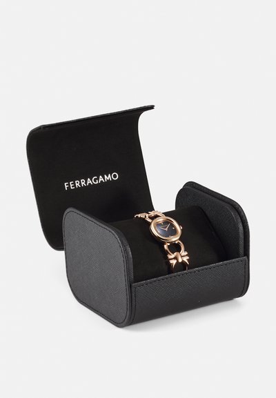 Rose gold watch with a round face and black dial, placed on a soft black cushion within a black box featuring the Ferragamo logo.